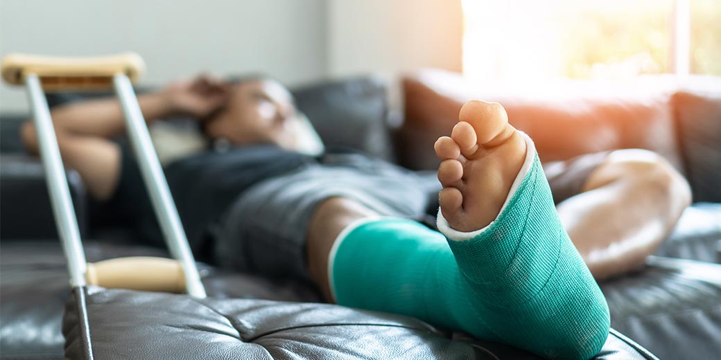 Mann liegt mit eingegipstem Fuß und Krücke auf dem Sofa