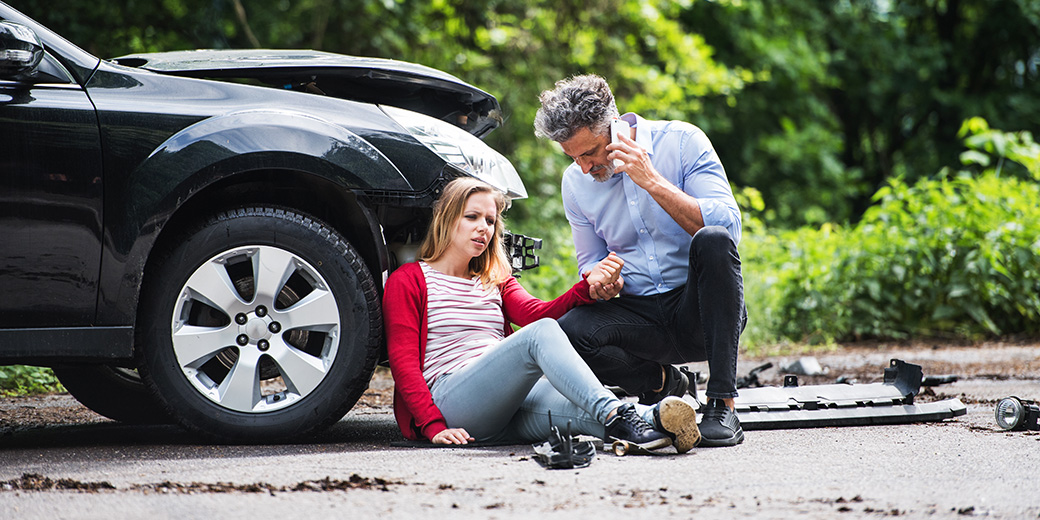 Eine verletzte Frau lehnt sich an ihr Auto und bekommt Hilfe von einem Mann